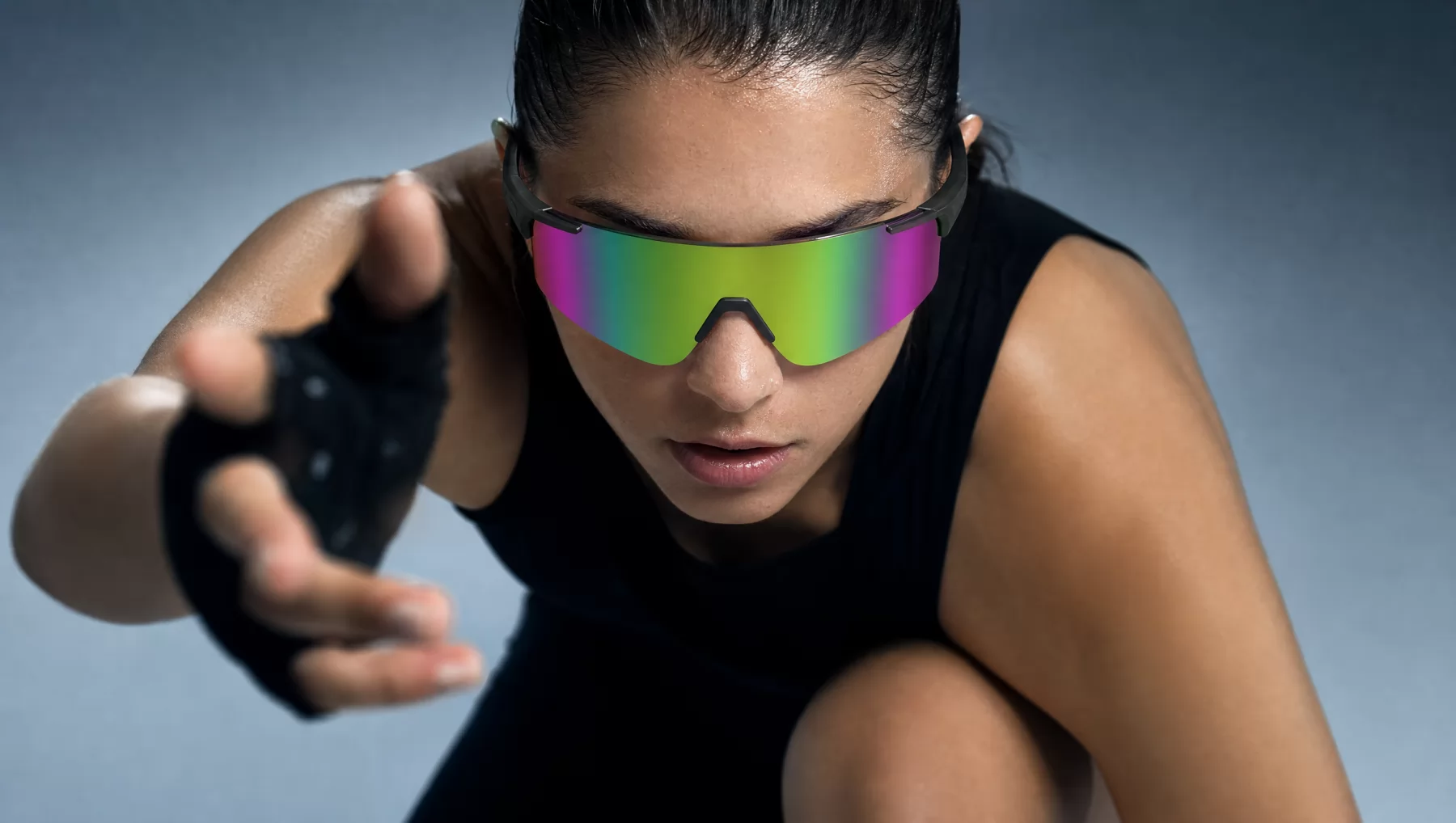 An athlete crouched forward wearing wraparound sport sunglasses with iridescent mirrored lenses, one hand reaching toward the camera